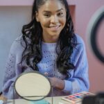 Positive African American female blogger sitting at table with cosmetics and mirror and shooting video on mobile phone